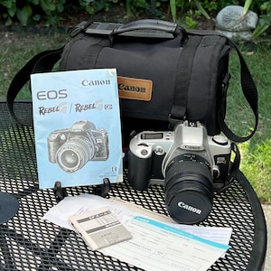 May include: A silver and black Canon EOS Rebel G camera with a lens, a black camera bag with a Canon logo patch, an instruction manual featuring a camera illustration, and several paper documents including a warranty card and lens guide.