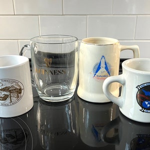 May include: Four mugs, two white ceramic, one clear glass, and one white ceramic with a gold rim. The mugs feature various designs, including a village scene, a space shuttle, and a military insignia. The mugs are on a black surface.