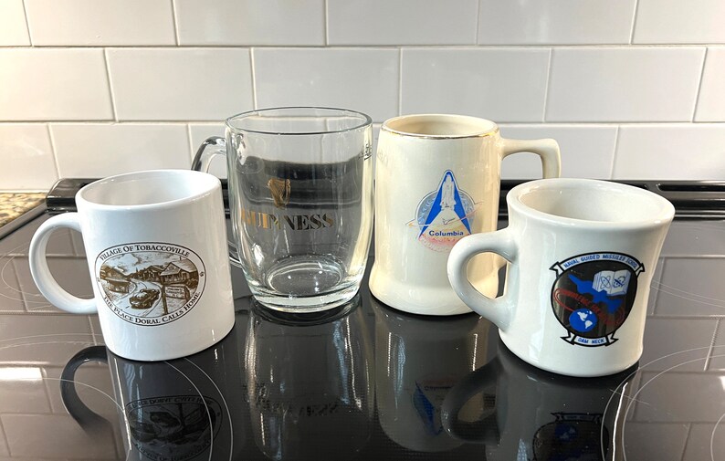 May include: Four mugs, two white ceramic, one clear glass, and one white ceramic with a gold rim. The mugs feature various designs, including a village scene, a space shuttle, and a military insignia. The mugs are on a black surface.