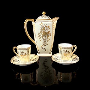 May include: A porcelain coffee set featuring a tall, square-shaped coffee pot with a lid and two matching cups with saucers. The set is cream-colored with gold floral patterns, gold trim on the edges, and gold handles on the pot and cups.