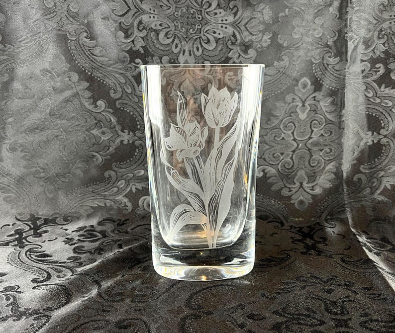 May include: A clear glass vase featuring an etched design of two tulips with detailed petals and leaves. The cylindrical vessel has a thick, solid base and smooth, transparent walls, showcasing the intricate floral engraving on the front surface.