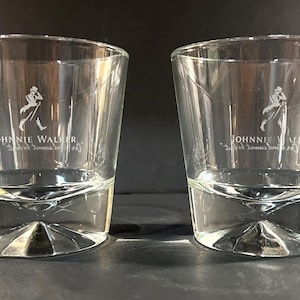 May include: Two clear, lowball whiskey glasses. Each glass features the "Johnnie Walker" logo, a striding man, etched in white. The glasses have a unique, faceted base design and a slightly flared rim. Ideal for serving spirits.