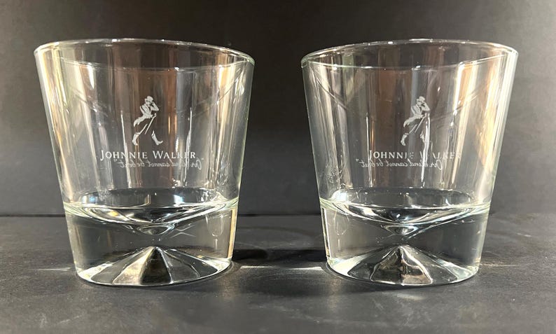 May include: Two clear, lowball whiskey glasses. Each glass features the "Johnnie Walker" logo, a striding man, etched in white. The glasses have a unique, faceted base design and a slightly flared rim. Ideal for serving spirits.