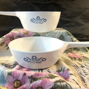May include: Two white ceramic bowls with blue floral designs and handles. The bowls are on a floral patterned fabric.