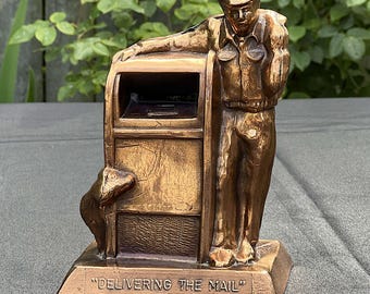 Op de afbeelding: Bronzen sculptuur van een postbode die tegen een brievenbus leunt, met een hond aan de voet. De sculptuur is gegraveerd met de woorden "DELIVERING THE MAIL". De figuur draagt een uniform en pet.