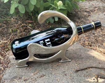 May include: A silver metal wine bottle holder shaped like a stylized bird with a grapevine design. The holder is holding a bottle of red wine.