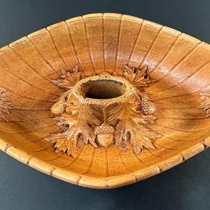 Puede incluir: Una fuente de madera en forma de diamante con un agujero central. La fuente presenta diseños tallados de hojas y bellotas alrededor del agujero y en los lados. La madera tiene un tono marrón cálido y parece estar en buen estado.