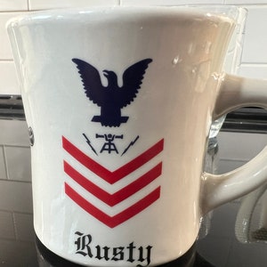 May include: White ceramic mug featuring a blue eagle, red and white stripes, and the text "Rusty".