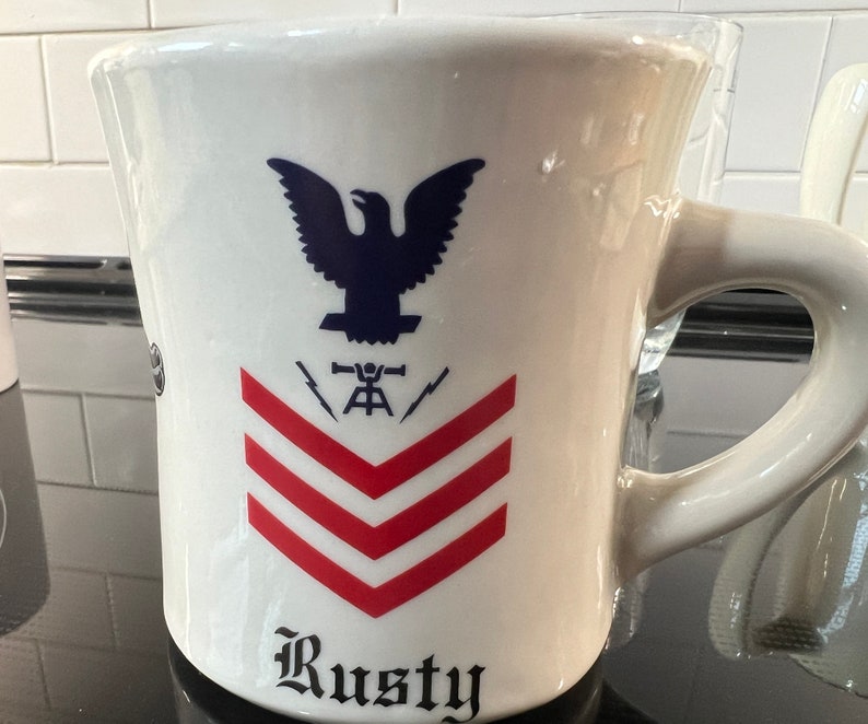 May include: White ceramic mug featuring a blue eagle, red and white stripes, and the text "Rusty".