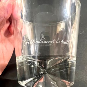 May include: A clear, cylindrical glass with a geometric base, partially filled with a clear liquid. The glass features the text "Our blend cannot be beat" and a logo. The glass is held by a hand.