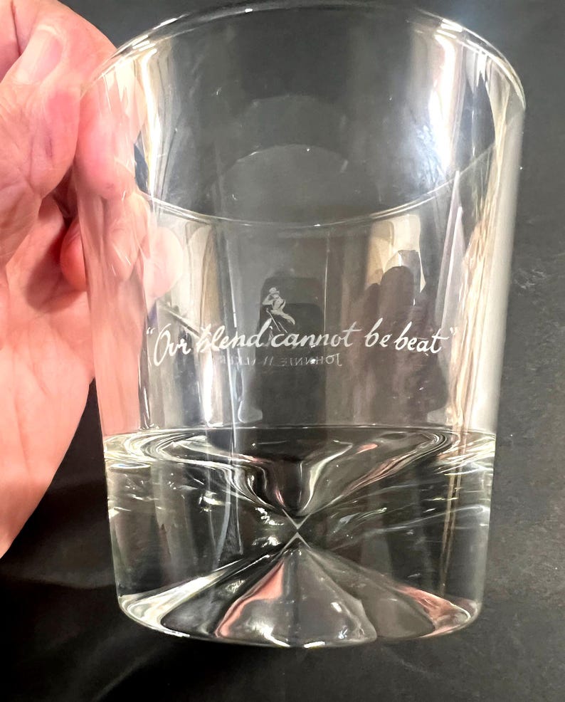 May include: A clear, cylindrical glass with a geometric base, partially filled with a clear liquid. The glass features the text "Our blend cannot be beat" and a logo. The glass is held by a hand.