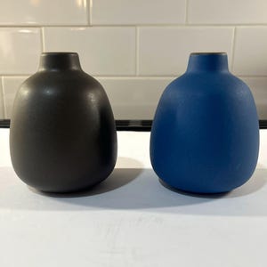 May include: Two small, rounded vases with narrow necks. One vase is matte black, and the other is matte blue. Both items feature a smooth, solid-colored finish with a bulbous body shape, standing side by side on a flat, white surface.