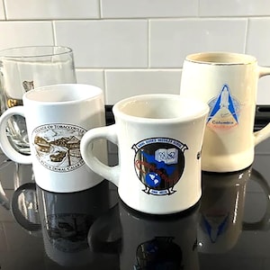 May include: Four ceramic mugs with different designs. One mug is clear glass. The mugs are on a black surface. The designs include a village scene, a school logo, and a religious image.