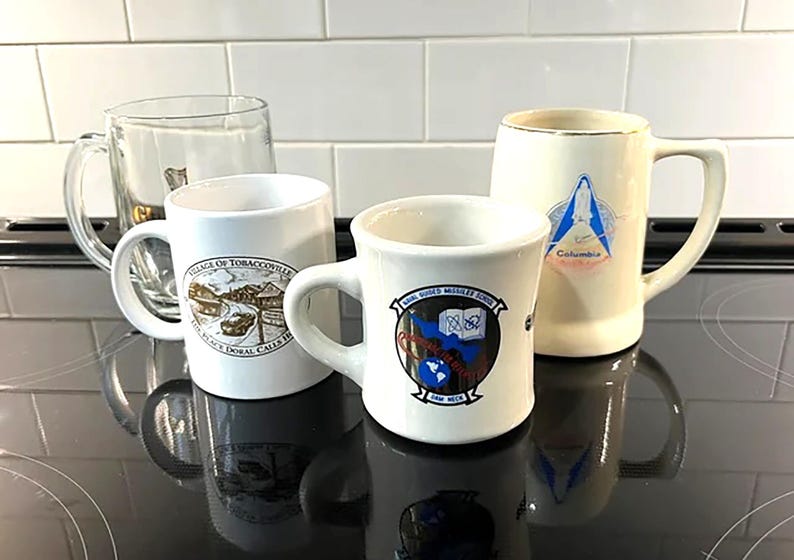 May include: Four ceramic mugs with different designs. One mug is clear glass. The mugs are on a black surface. The designs include a village scene, a school logo, and a religious image.