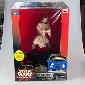 May include: A Star Wars Episode I Qui-Gon Jinn interactive talking bank in its original packaging. The figure is in a fighting stance, holding a lightsaber. The box is red and black with the Star Wars logo and "Try Me!" text.
