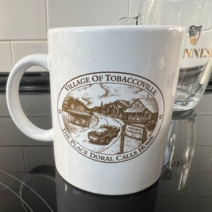 May include: White ceramic coffee mug with a brown illustration of a town with the text "Village of Tobaccoville The Place Doral Calls Home" on the side.