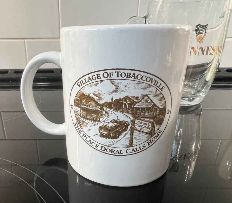 May include: White ceramic coffee mug with a brown illustration of a town with the text "Village of Tobaccoville The Place Doral Calls Home" on the side.