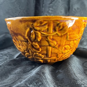 May include: A round ceramic bowl with a glossy, honey-brown glaze. The exterior features a raised, embossed pattern of woven basketry textures intertwined with detailed leaves and vines, creating a textured surface across the entire outer wall.