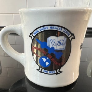 May include: White ceramic coffee mug with a blue and gold logo that reads "Naval Guided Missiles School" and "Dam Neck".