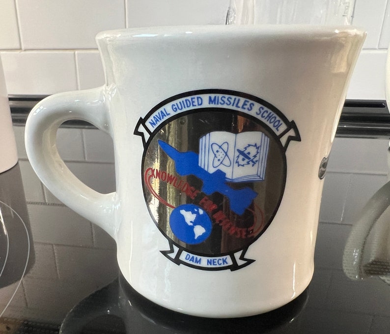 May include: White ceramic coffee mug with a blue and gold logo that reads "Naval Guided Missiles School" and "Dam Neck".