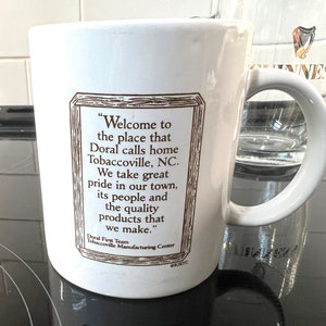 May include: White ceramic mug with a brown framed quote that reads "Welcome to the place that Doral calls home Tobaccoville, NC. We take great pride in our town, its people and the quality products that we make." The quote is attributed to "Doral First Team Tobaccoville Manufacturing Center".