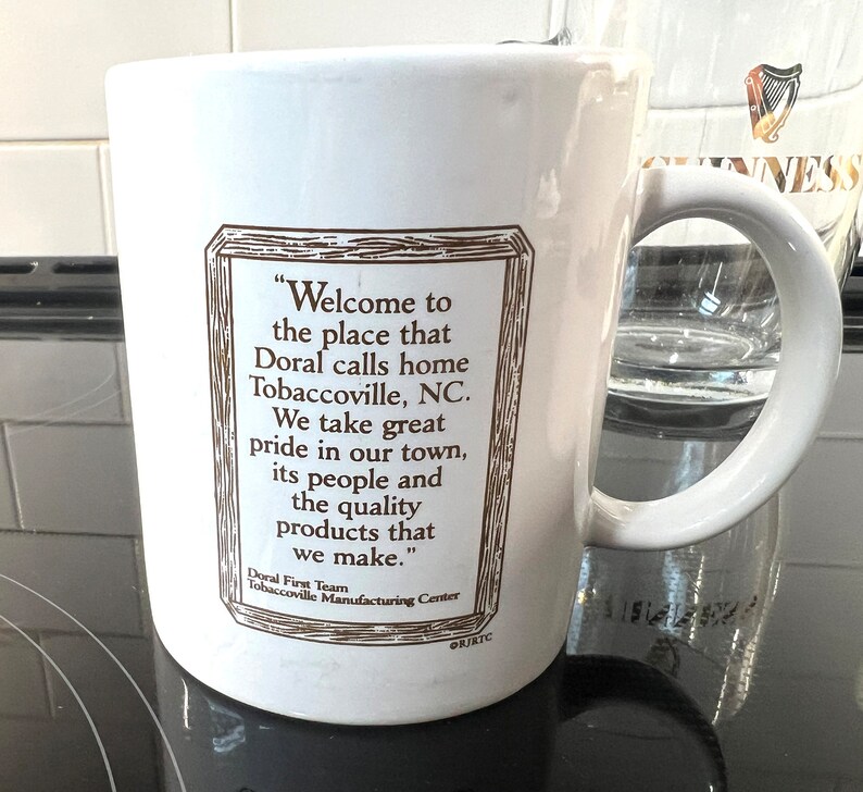 May include: White ceramic mug with a brown framed quote that reads "Welcome to the place that Doral calls home Tobaccoville, NC. We take great pride in our town, its people and the quality products that we make." The quote is attributed to "Doral First Team Tobaccoville Manufacturing Center".