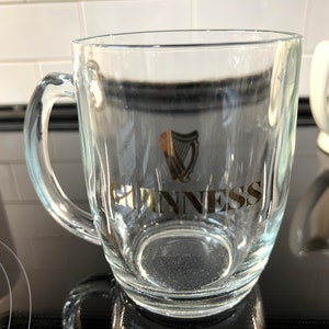 May include: A clear glass pint glass with a gold Guinness logo and harp design. The glass has a handle and is sitting on a black surface.