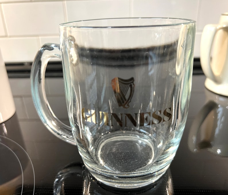 May include: A clear glass pint glass with a gold Guinness logo and harp design. The glass has a handle and is sitting on a black surface.