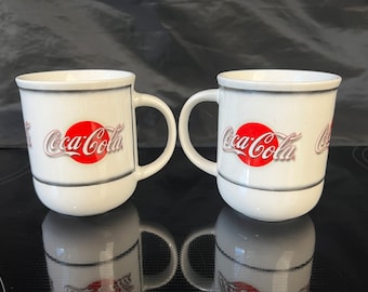 May include: Two white ceramic mugs with a red Coca-Cola logo and silver trim. The mugs have a handle on the right side.