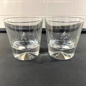 May include: Two clear, lowball whiskey glasses with a faceted base. Each glass has the "Johnnie Walker" logo in white. The glasses are partially filled with a clear liquid, likely a beverage, and sit on a dark surface.