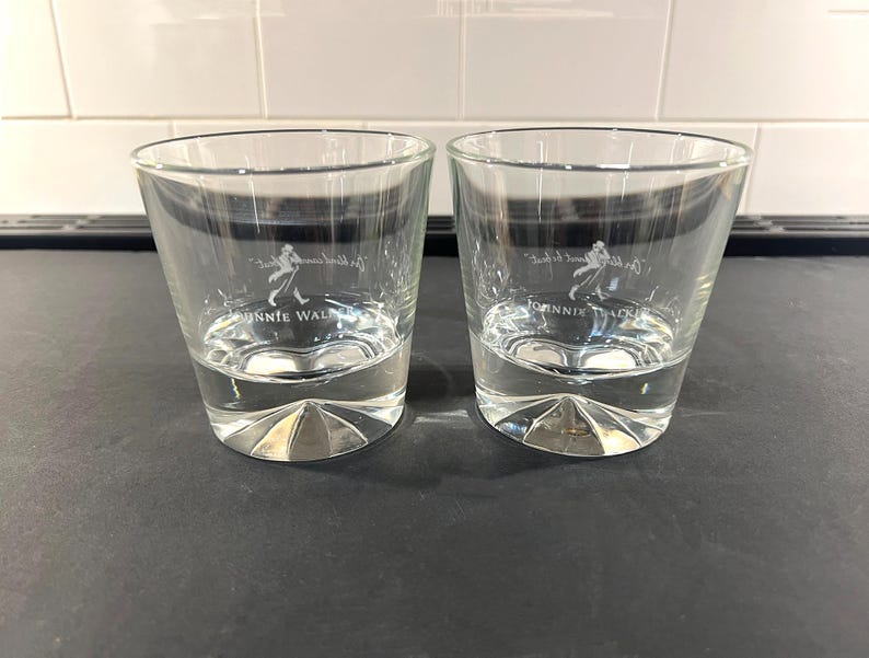 May include: Two clear, lowball whiskey glasses with a faceted base. Each glass has the "Johnnie Walker" logo in white. The glasses are partially filled with a clear liquid, likely a beverage, and sit on a dark surface.