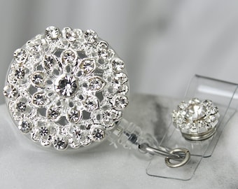 May include: A silver rhinestone badge reel with a clear plastic strap and a smaller matching rhinestone accent. The badge reel features a floral design with multiple clear rhinestones. The background is a soft, blurred white.
