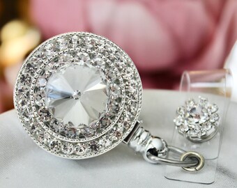 May include: A sparkling, silver badge reel with a large, round, clear crystal center surrounded by multiple rows of smaller crystals. A smaller, matching crystal accent is attached to a clear plastic holder. The background is a soft, blurred pink and white.
