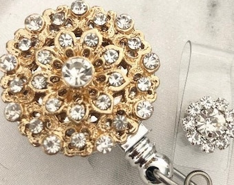 May include: A gold-toned badge reel with a large, round, crystal-studded design. A smaller, matching crystal accent is attached to a clear plastic clip. The reel has a silver-colored metal clip and a retractable cord.