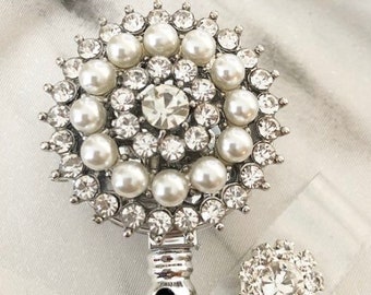 May include: A silver badge reel with a round, ornate design. The reel features a central clear rhinestone surrounded by faux pearls and smaller clear rhinestones. A smaller matching button is also included.