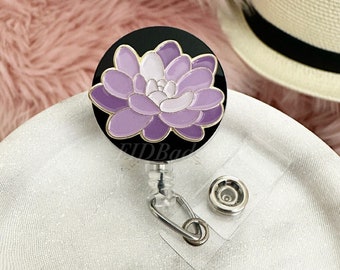 May include: A purple flower with white accents on a black background, with a gold rim, is attached to a retractable badge reel with a clear plastic clip.