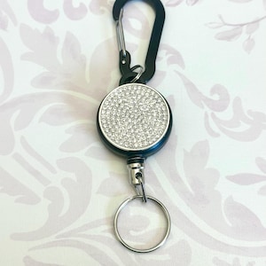 May include: A black retractable badge holder with a silver carabiner clip and a silver key ring. The round holder is black with a silver rhinestone-covered face.