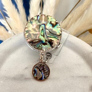May include: A round badge reel with a shimmering abalone shell design. The badge reel has a gold-colored trim and a smaller, matching charm hanging below. The iridescent colors of the shell include blues, greens, and browns.