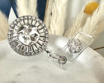 May include: A sparkling badge reel with a large, round, clear rhinestone centerpiece surrounded by smaller rhinestones. A smaller, matching rhinestone accent is attached to the clear plastic reel clip. The badge reel is silver-toned.