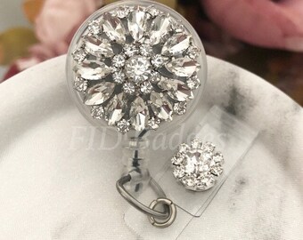 May include: A clear, round badge reel with a sparkling, crystal-like floral design. The badge reel has a silver-colored metal clip and a smaller matching charm. The background is a soft, blurred pink and white.