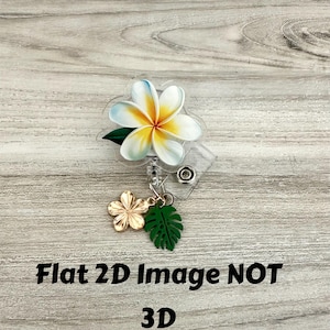 K&ouml;nnte beinhalten: Ein durchsichtiges Ausweis-Jojo mit einem Design einer wei&szlig;en und gelben Plumeria-Blume mit einem gr&uuml;nen Blatt. Ein goldener Blumen- und gr&uuml;ner Blatt-Anh&auml;nger h&auml;ngt darunter. Der Text "Flat 2D Image NOT 3D" ist in Schwarz gedruckt.