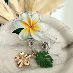 K&ouml;nnte beinhalten: Ein durchsichtiges Ausweis-Jojo mit floralem Design. Das Abzeichen zeigt eine wei&szlig;e und gelbe Plumeria-Blume mit blauen Akzenten und einem gr&uuml;nen Blatt. Ein goldener Blumen- und gr&uuml;ner Blatt-Anh&auml;nger baumelt an der Rolle.