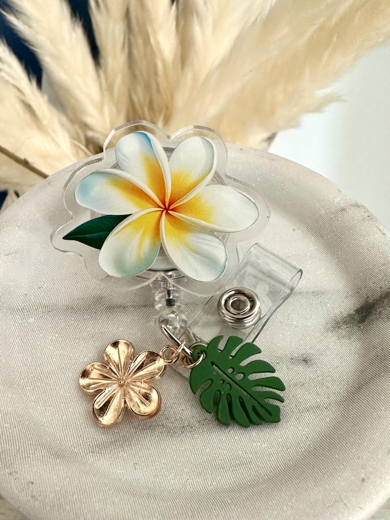 K&ouml;nnte beinhalten: Ein durchsichtiges Ausweis-Jojo mit floralem Design. Das Abzeichen zeigt eine wei&szlig;e und gelbe Plumeria-Blume mit blauen Akzenten und einem gr&uuml;nen Blatt. Ein goldener Blumen- und gr&uuml;ner Blatt-Anh&auml;nger baumelt an der Rolle.