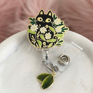 May include: A black cat badge reel with a floral design. The cat is surrounded by white daisies and green leaves, with gold accents. The reel has a clear plastic retractable cord and a leaf charm.