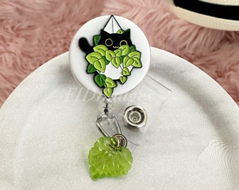 May include: A black cat with green leaves around it is featured on a white round badge with a clear retractable badge holder and a green leaf charm.