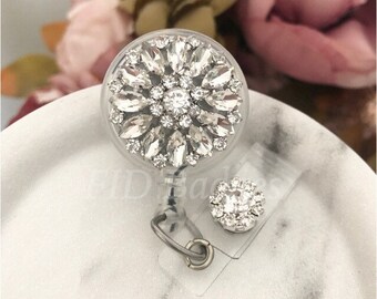 May include: A clear plastic retractable badge holder with a large rhinestone flower design and a smaller rhinestone accent. The holder is on a white surface.