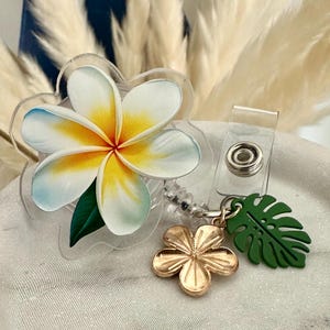 K&ouml;nnte beinhalten: Ein durchsichtiges Acryl-Ausweis-Jojo mit einem Design einer wei&szlig;en, gelben und blauen Plumeria-Blume. Ein goldfarbener Blumenanh&auml;nger und ein gr&uuml;ner Bl&auml;tteranh&auml;nger h&auml;ngen an der Rolle, die einen transparenten Kunststoffclip hat.