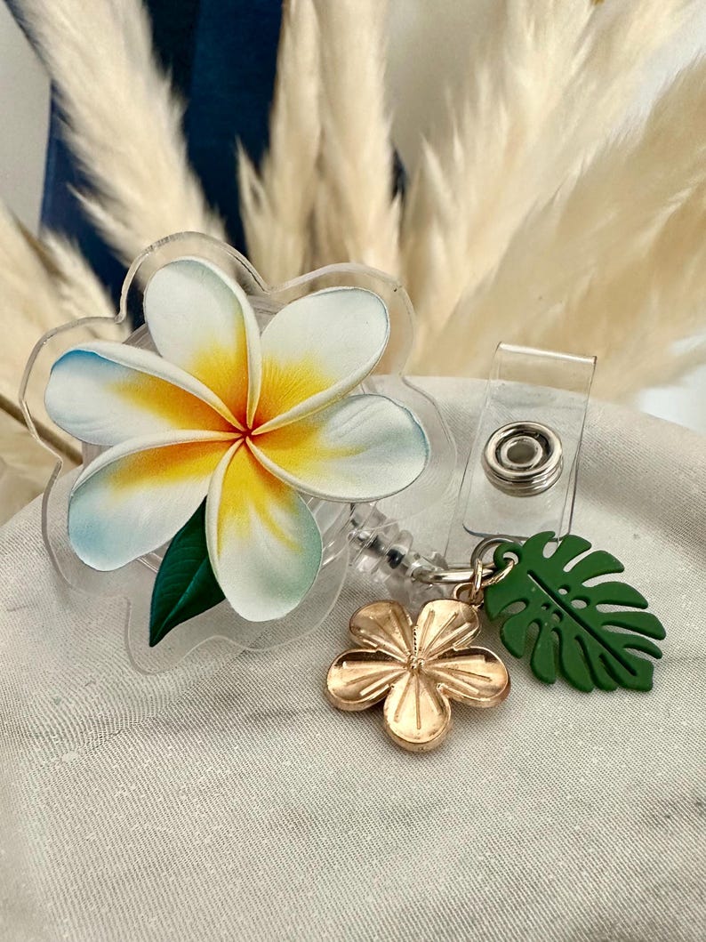K&ouml;nnte beinhalten: Ein durchsichtiges Acryl-Ausweis-Jojo mit einem Design einer wei&szlig;en, gelben und blauen Plumeria-Blume. Ein goldfarbener Blumenanh&auml;nger und ein gr&uuml;ner Bl&auml;tteranh&auml;nger h&auml;ngen an der Rolle, die einen transparenten Kunststoffclip hat.