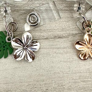 K&ouml;nnte beinhalten: Zwei einziehbare Ausweishalter mit Blumen- und Blattanh&auml;ngern. Ein Blumenanh&auml;nger ist silberfarben, der andere goldfarben. Jede Ausweisrolle hat einen gr&uuml;nen Blattanhänger und eine transparente Kunststoffrolle.
