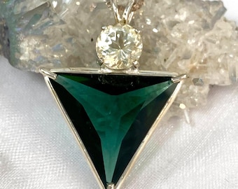 May include: A silver pendant necklace featuring a large, triangular, deep green gemstone set in a silver frame. Above the triangle sits a smaller, round, pale yellow gemstone. The pendant hangs from a thin, delicate silver chain.